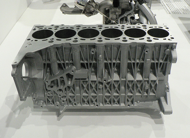 Auf einer wei&szlig;en Fl&auml;che ist ein metallischer Motorblock mit sechs Zylindern abgebildet. Die Struktur, die wahrscheinlich von einem Druckgusslieferanten in China hergestellt wurde, weist komplizierte geometrische Muster und verschiedene Befestigungspunkte auf und demonstriert ihre detaillierte Konstruktion. Im Hintergrund ist teilweise eine komplexe Rohranordnung zu erkennen