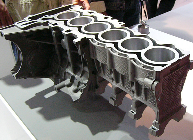 Eine Nahaufnahme eines geschnittenen Motorblocks, die die komplizierte innere Struktur und die Zylinderbohrungen zeigt. Das Metallteil liegt auf einem Tisch, mehrere Personen beobachten es im Hintergrund. Das Bild hebt die technischen und Designdetails des Motors hervor, mit freundlicher Genehmigung eines Aluminium-Druckgussunternehmens